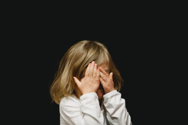 A little girl covering her face with her hands on a black background.