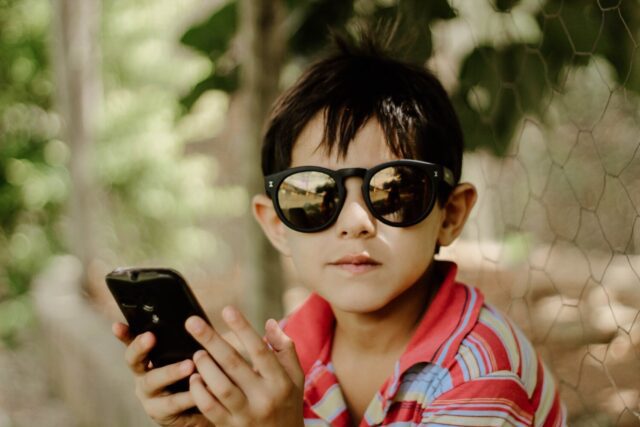 A boy wearing sunglasses and holding a cell phone.