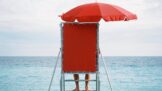 A person standing on a lifeguard stand with a red umbrella.