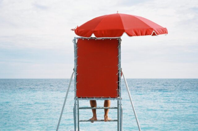 A person standing on a lifeguard stand with a red umbrella.