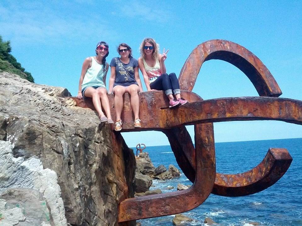 Three women sitting on top of a metal sculpture near the ocean.